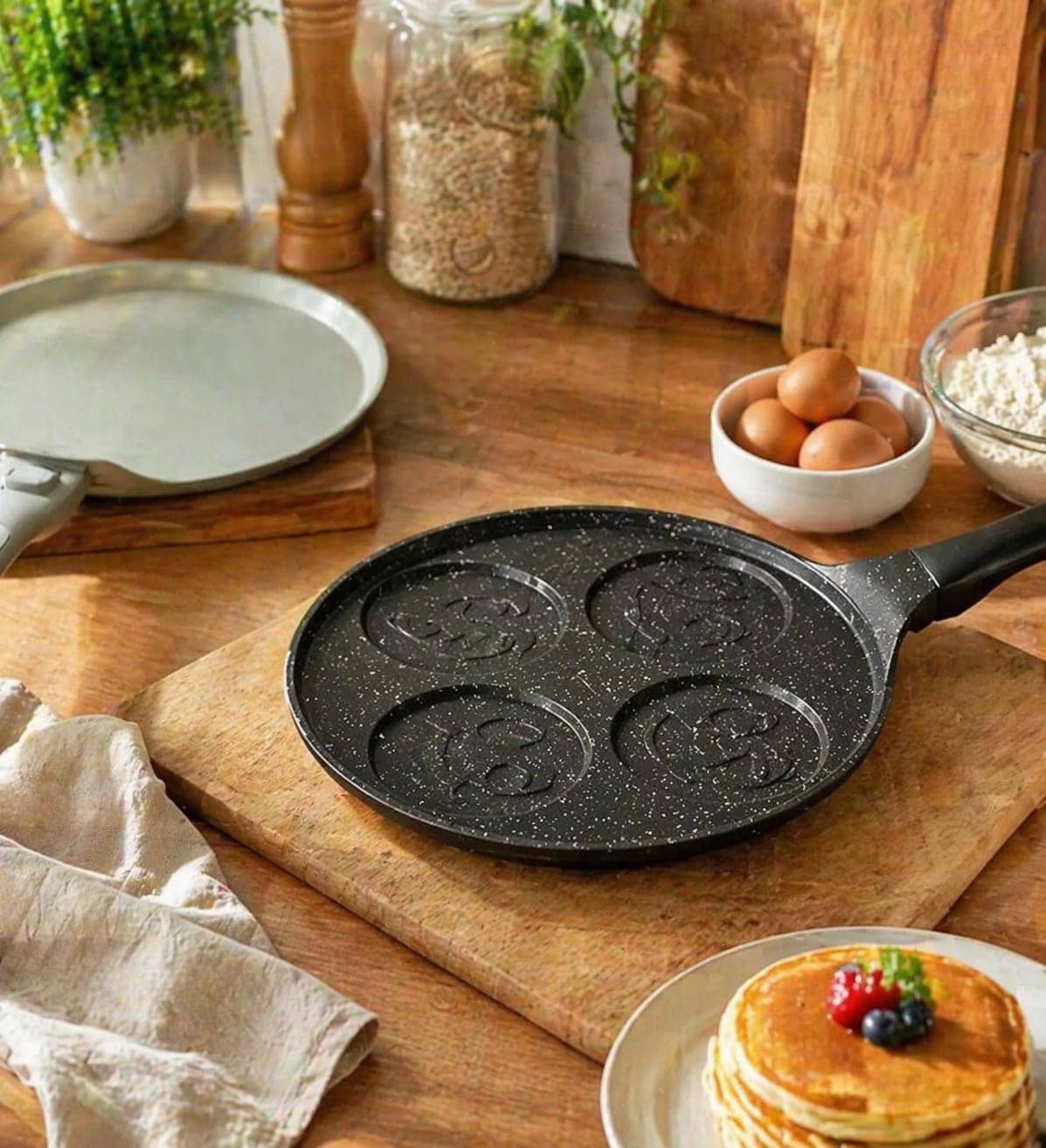 Une poêle noire antiadhésive avec quatre moules à motifs pour pancakes, posée sur une planche en bois. — vue 1